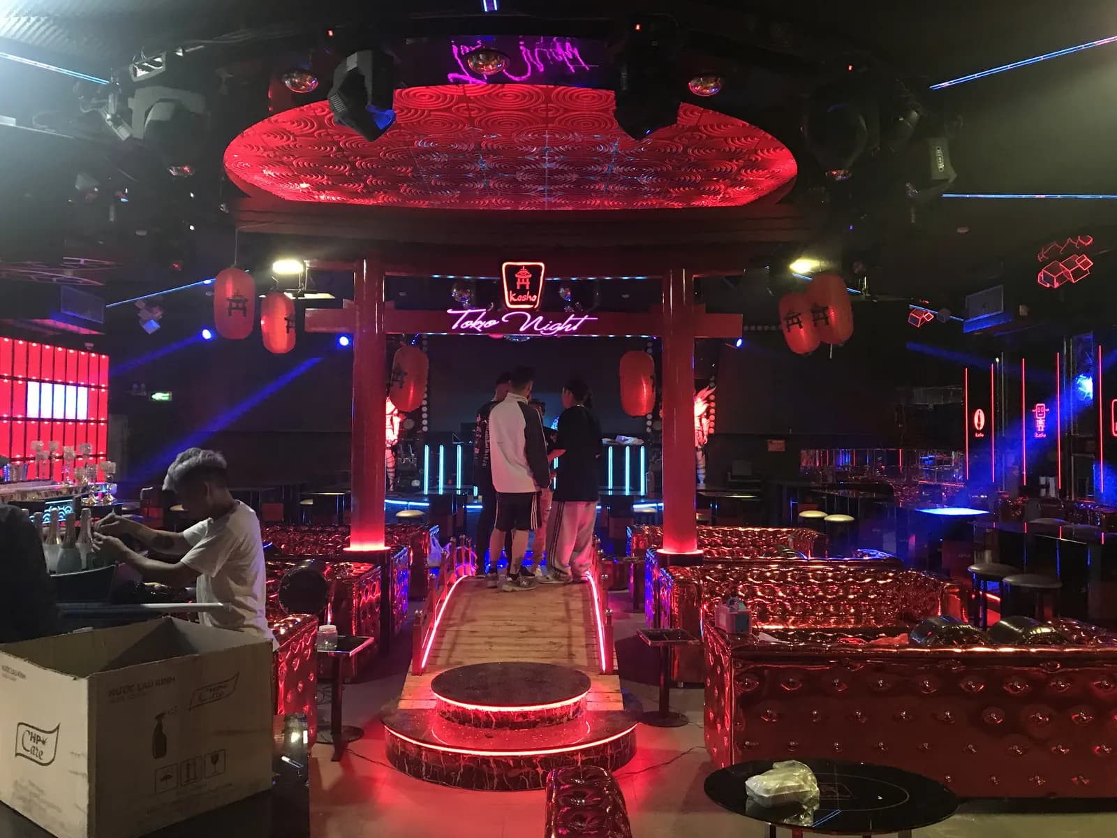 Góc nhìn khác của không gian club Kosho qua cổng torii đỏ, với đèn lồng trang trí, bàn DJ phía trong và các vùng ghế bọc kim loại đỏ xung quanh lối đi.