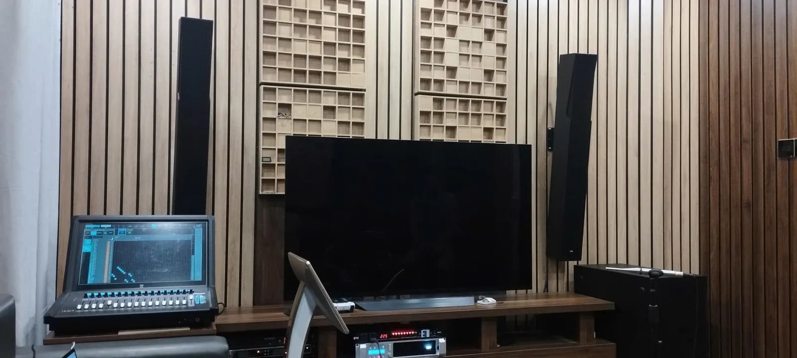 Phòng nghe - xem tại nhà riêng với loa đứng hai bên, subwoofer và bàn điều khiển số, vách gỗ tiêu âm dạng nan phía sau màn hình.